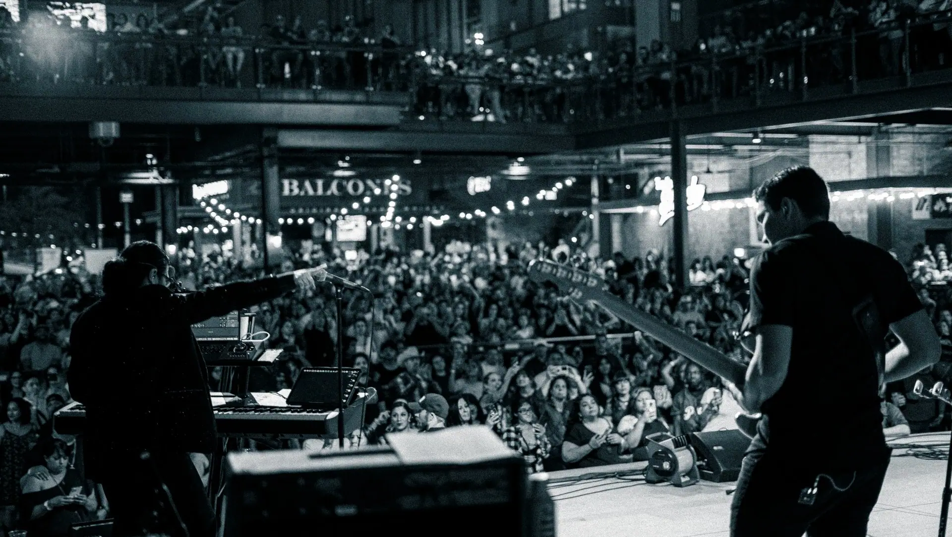 Black and white view from behind the band as a packed crowd fills a multi-level venue