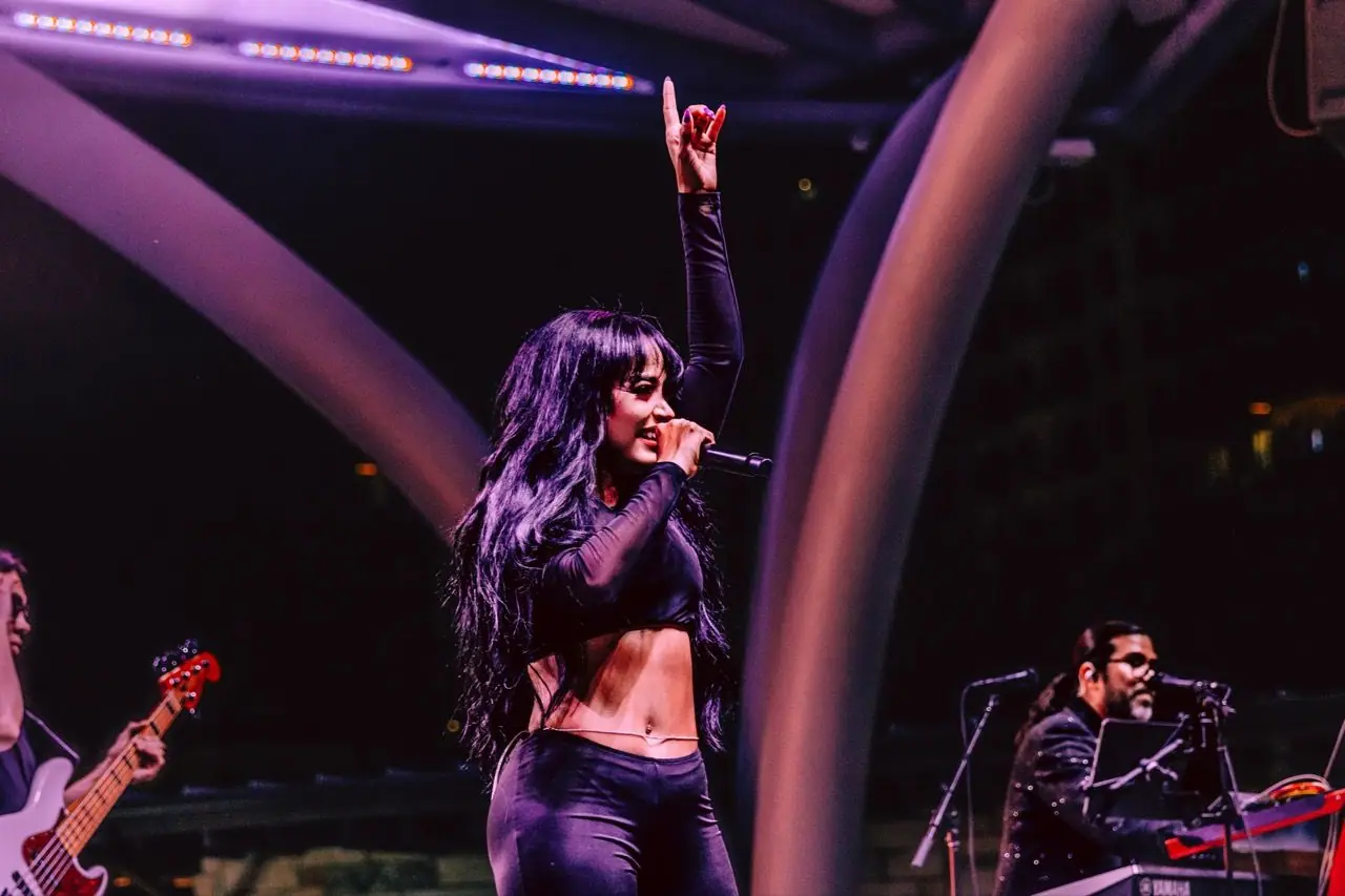 Carymel Rodríguez points skyward while singing on an outdoor stage beneath a curved canopy