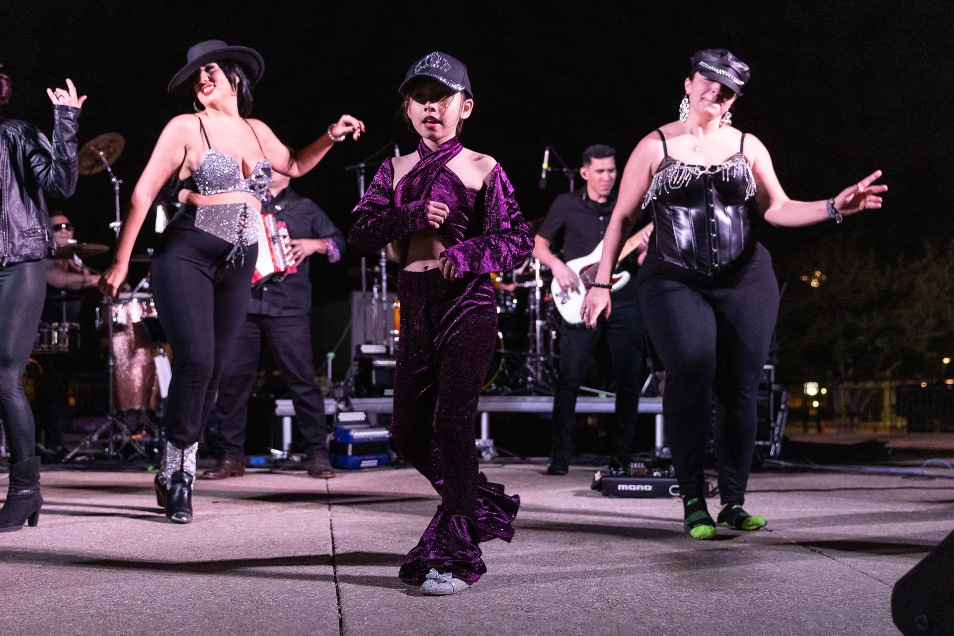 Three dancers in Selena-style costumes perform in front of the band on an outdoor stage