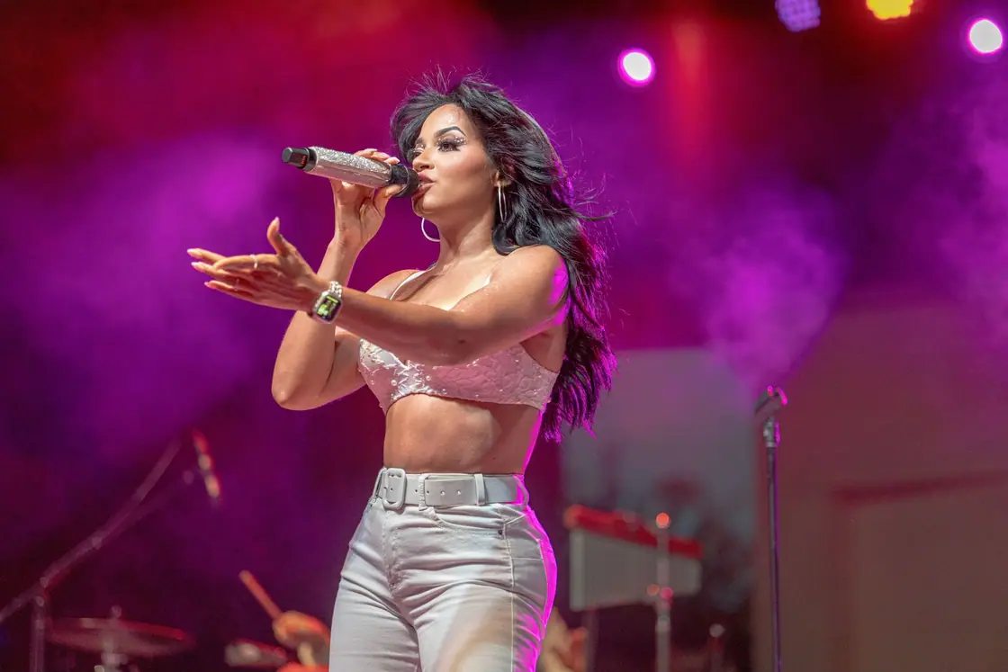 Carymel Rodríguez sings on stage in a sparkling crop top and white pants under pink and purple lights